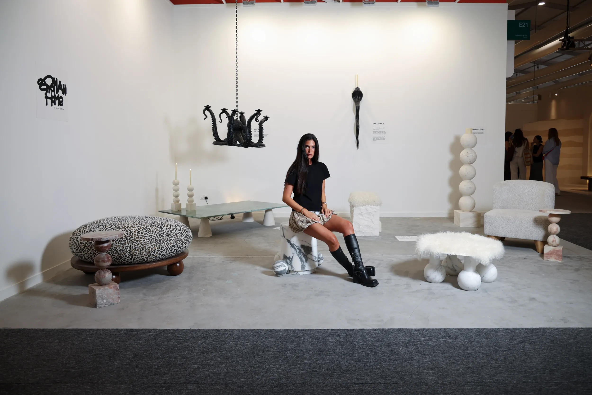 A seated woman surrounded by sculptural furniture and objects in a white exhibition space.