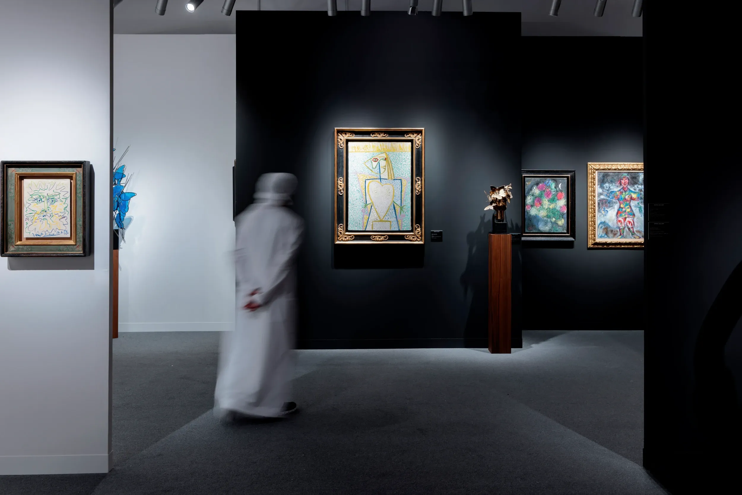 Visitor in a kandura walking past spotlit paintings in a Dubai gallery