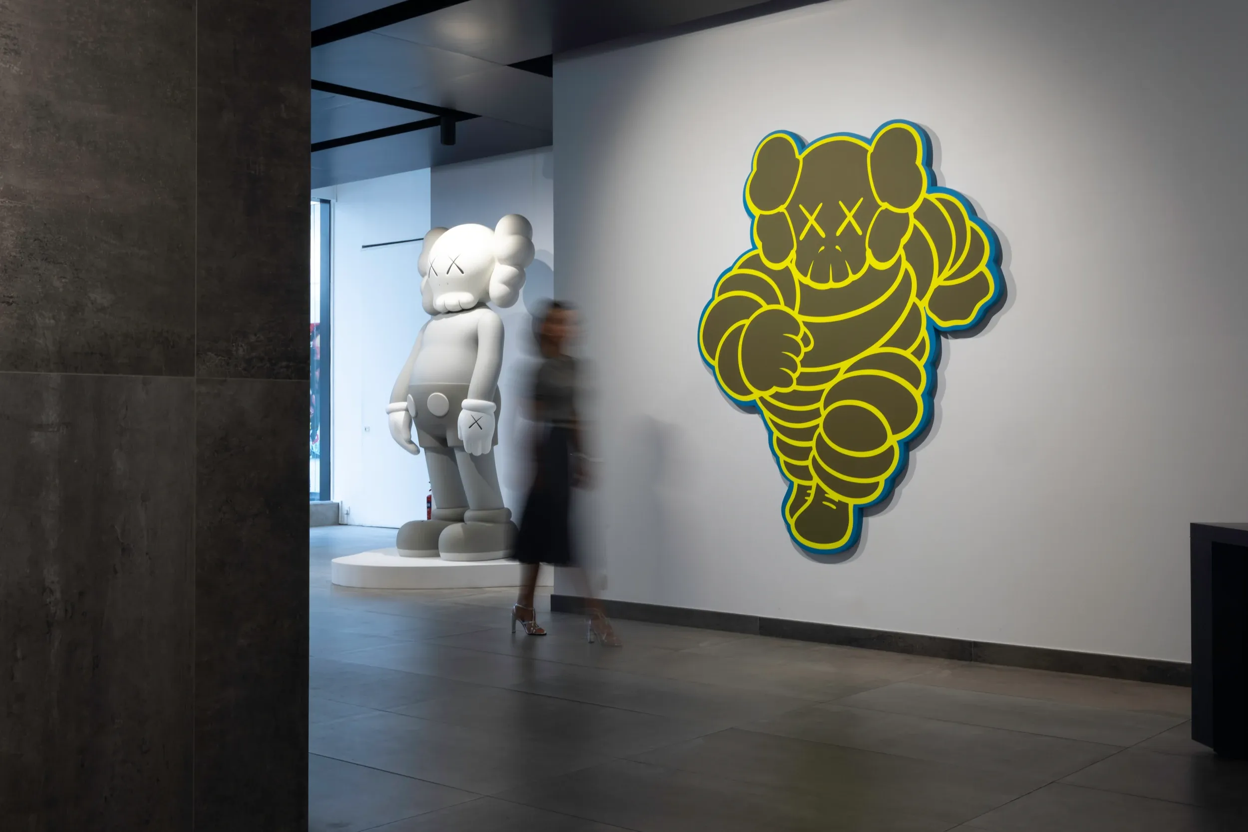 A visitor blurred in motion between a large grey sculpture and a black-and-yellow wall artwork.