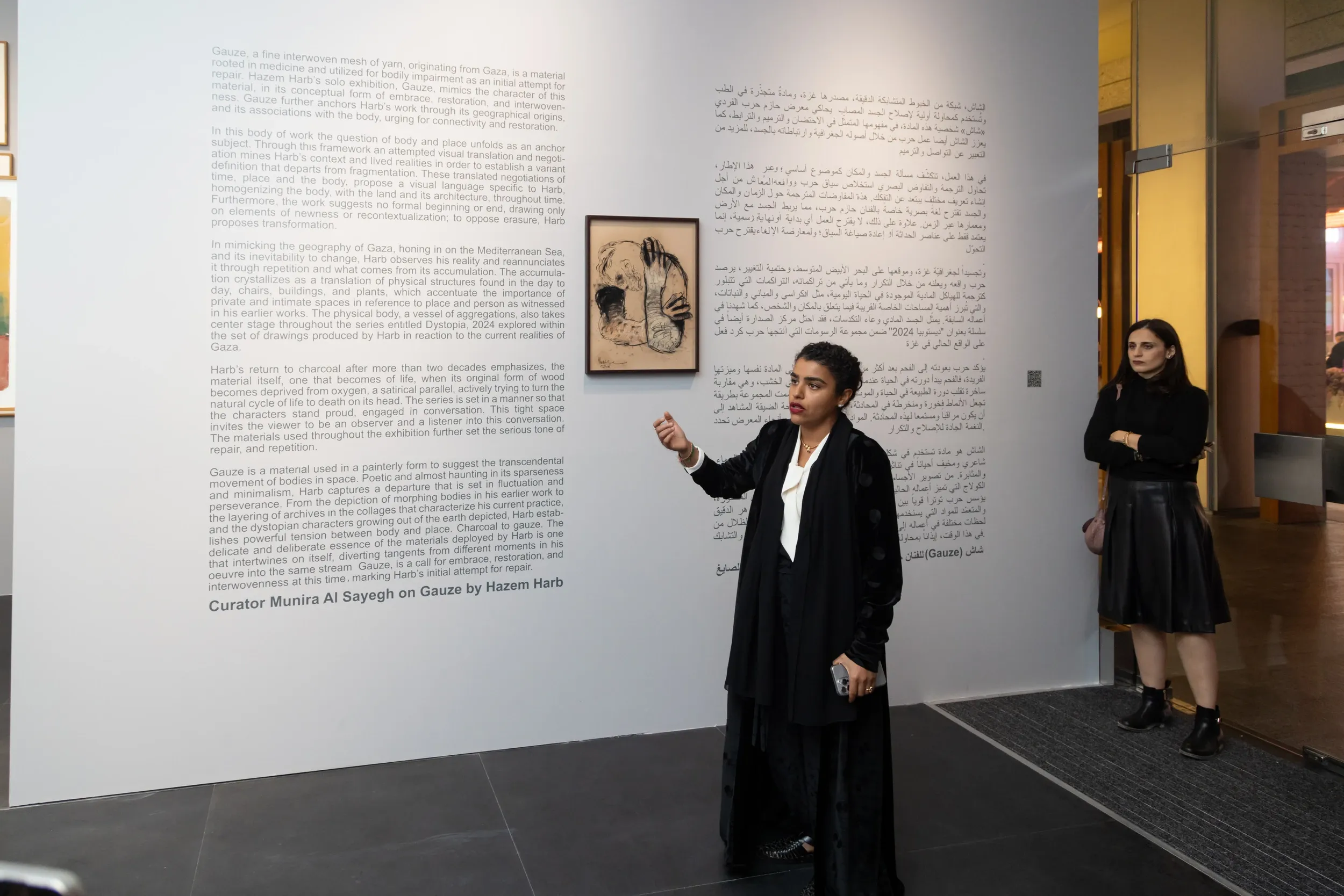 A woman gesturing beside exhibition wall text and framed artworks.