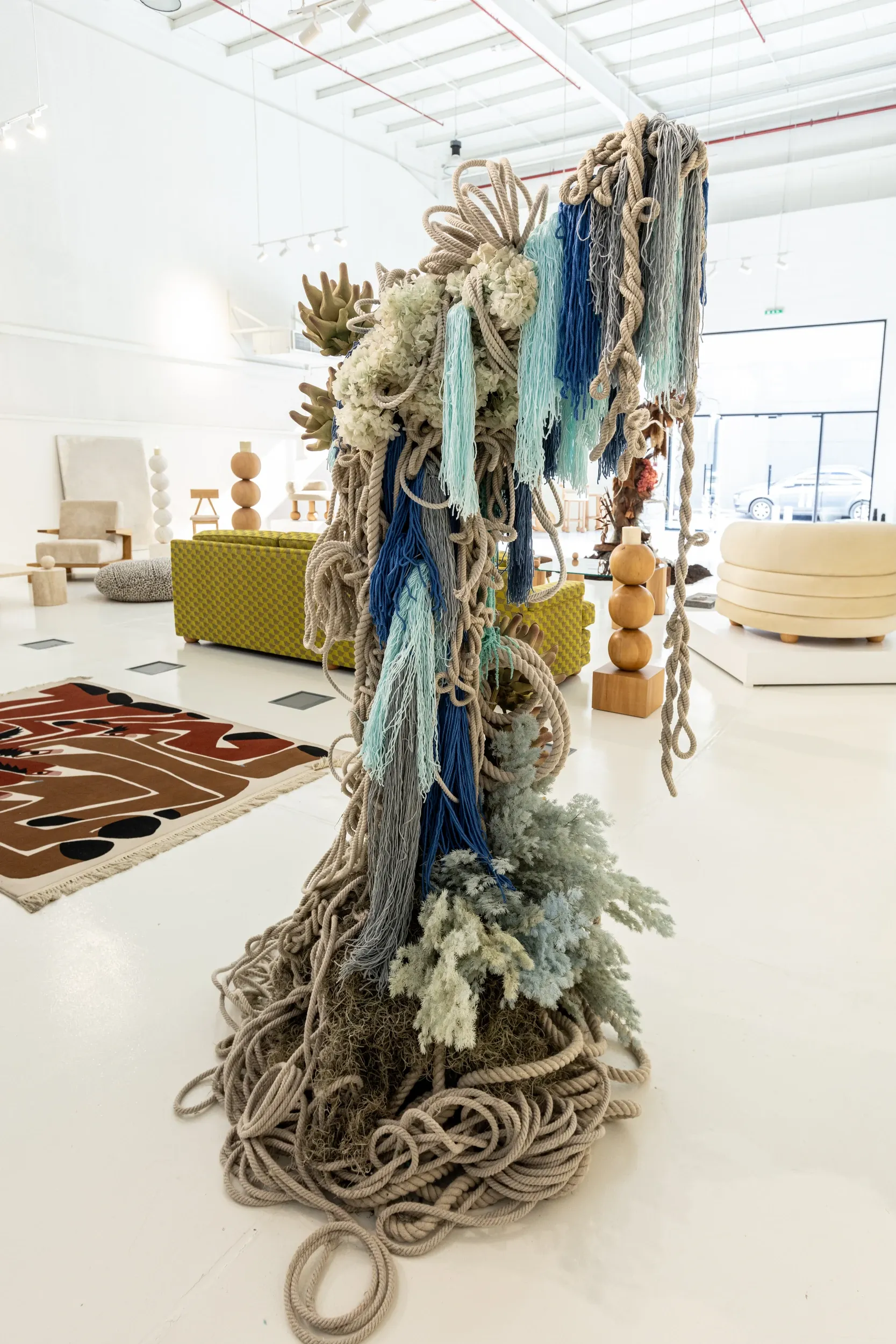 A tall textile sculpture made of hanging ropes and fibres in a bright exhibition space.