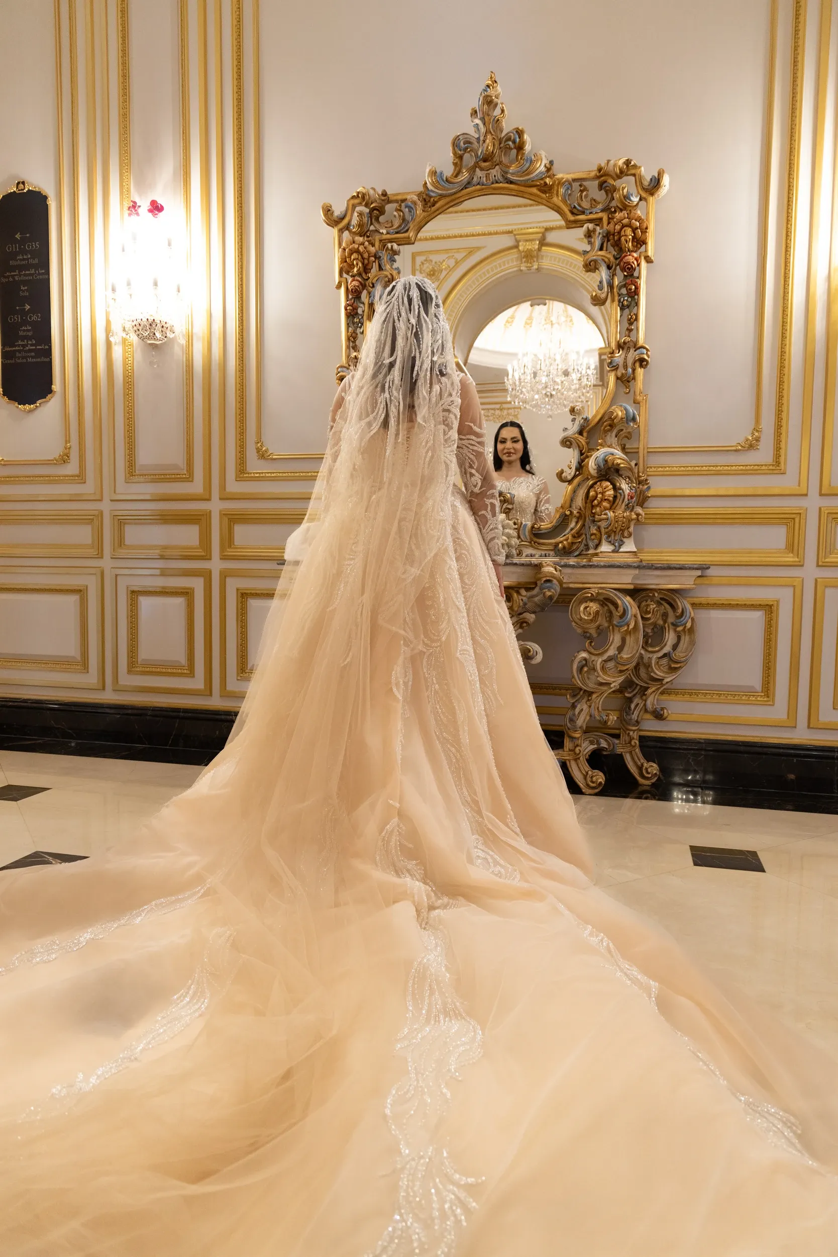 A bride's long lace train leading toward her reflection in an ornate gold mirror.