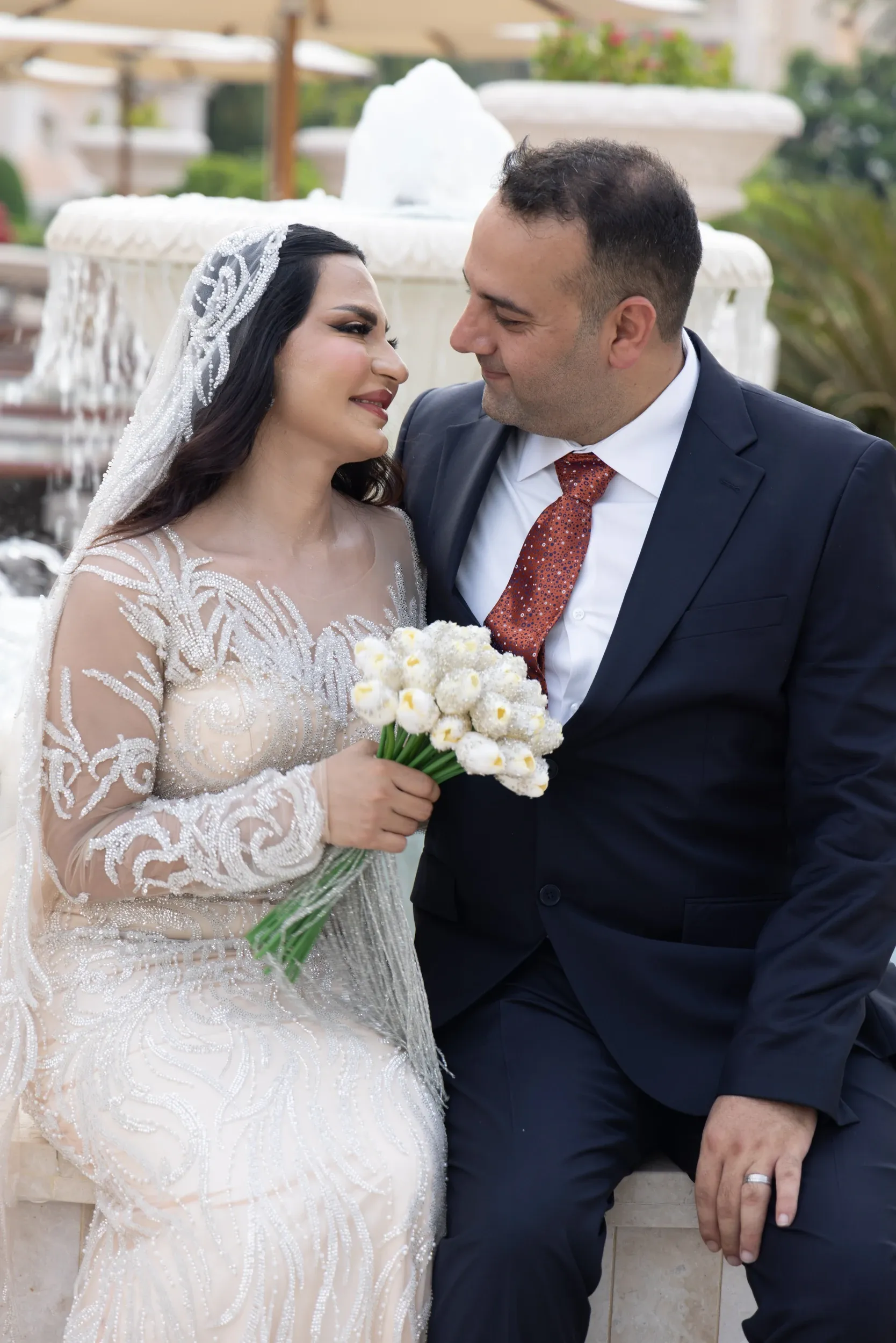 A seated wedding couple sharing a close moment beside a fountain.