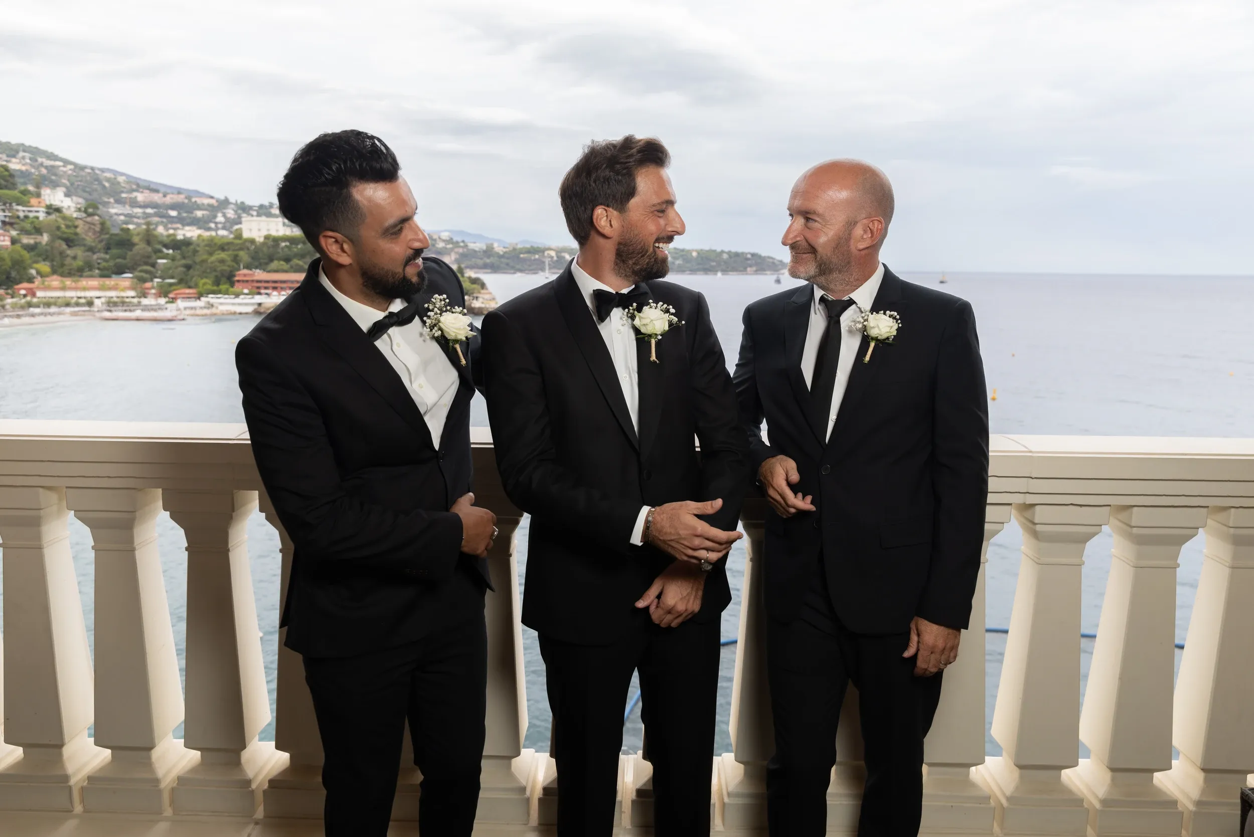 Three men in dark wedding suits talking on a sea-view balcony.