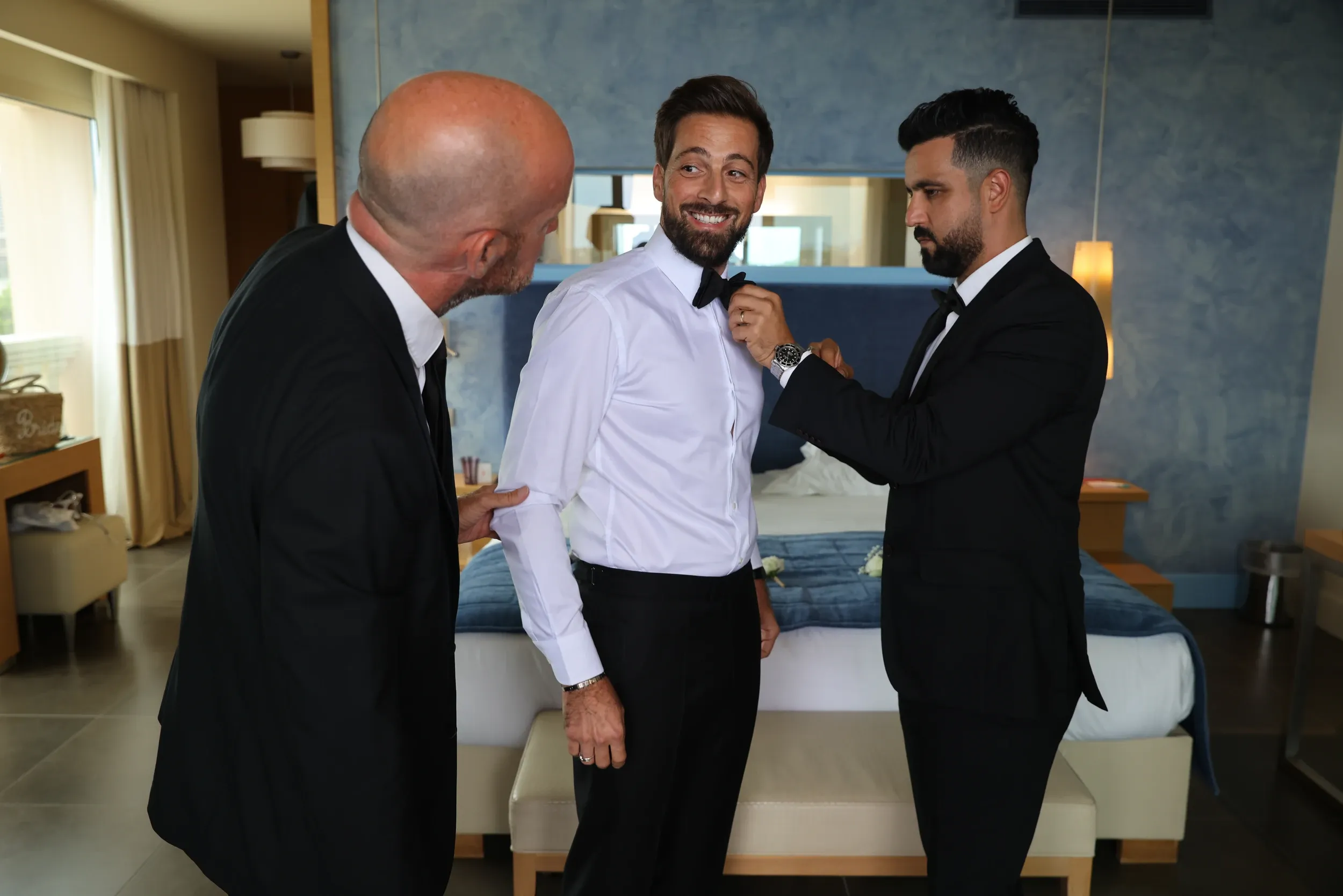 Two men helping a smiling groom adjust his bow tie in a hotel room.