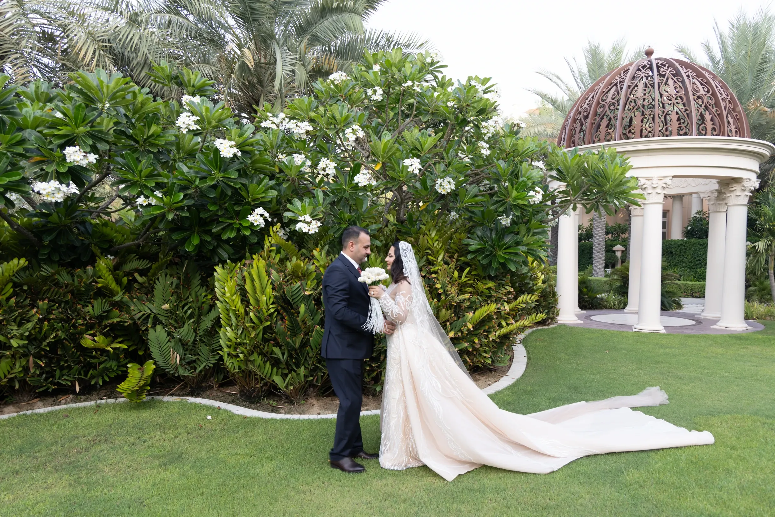 A wedding couple on a garden lawn beside a domed pavilion.