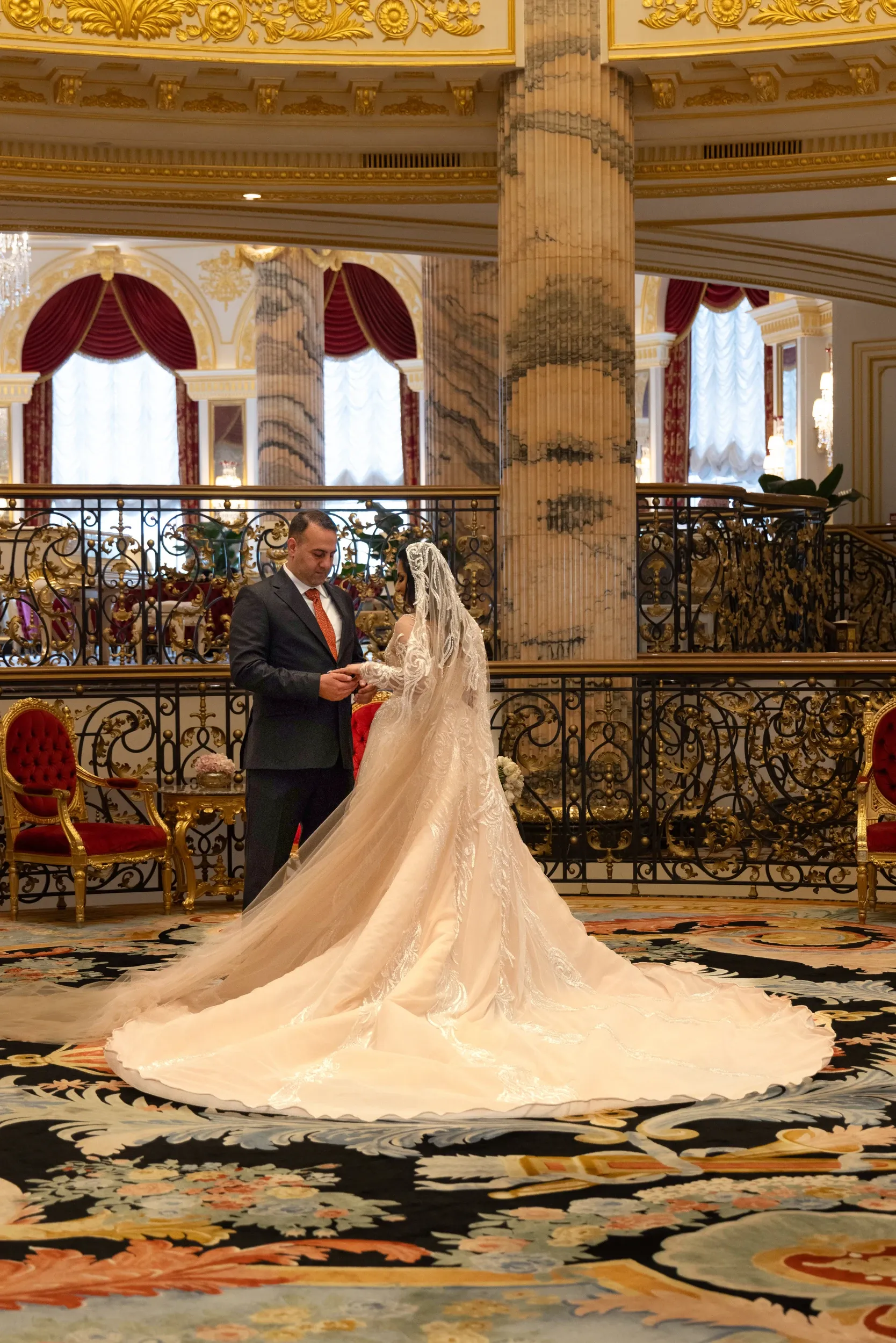 A wedding couple with a sweeping bridal train beside an ornate interior balustrade.