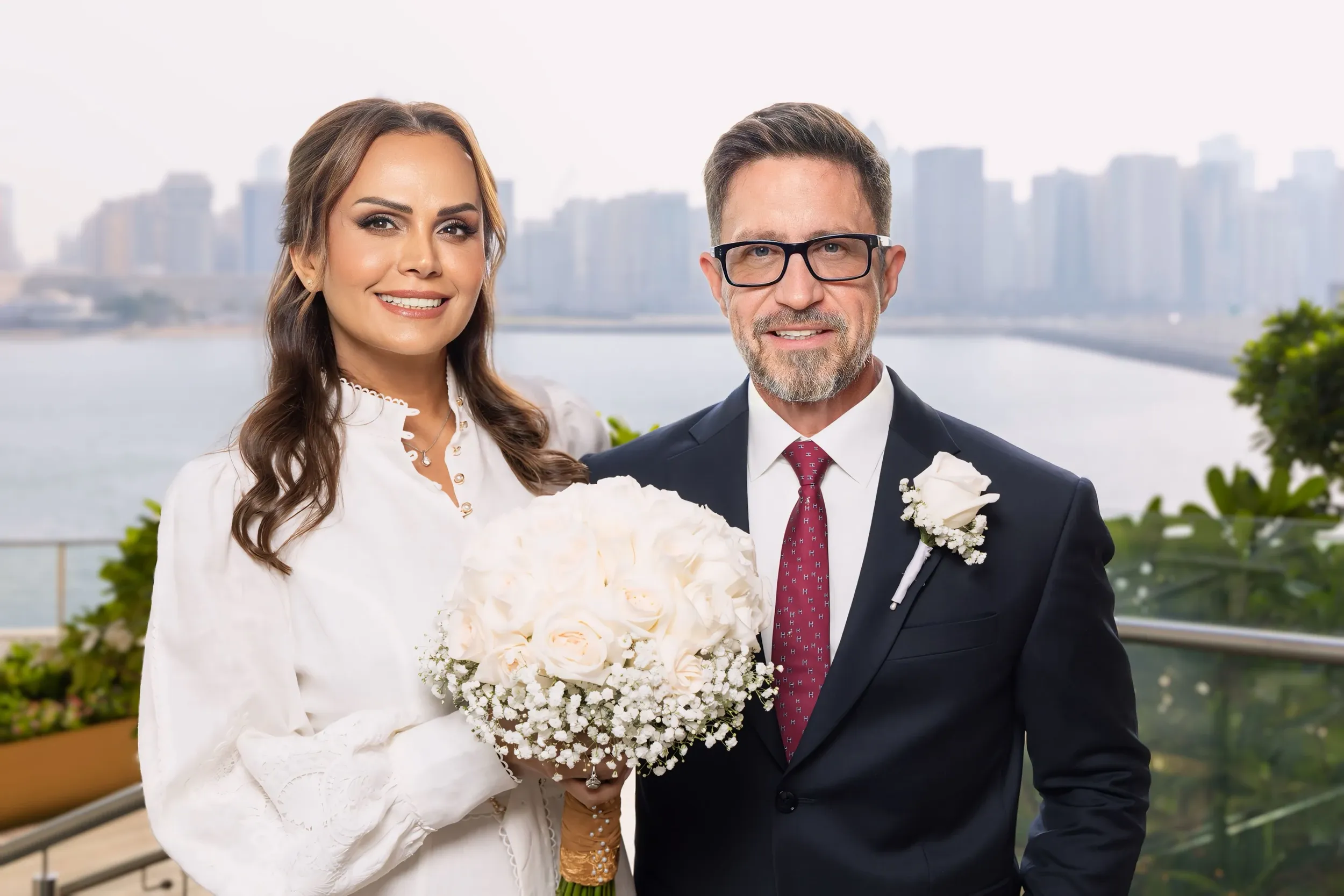 A wedding couple with a white bouquet and a waterfront skyline behind.