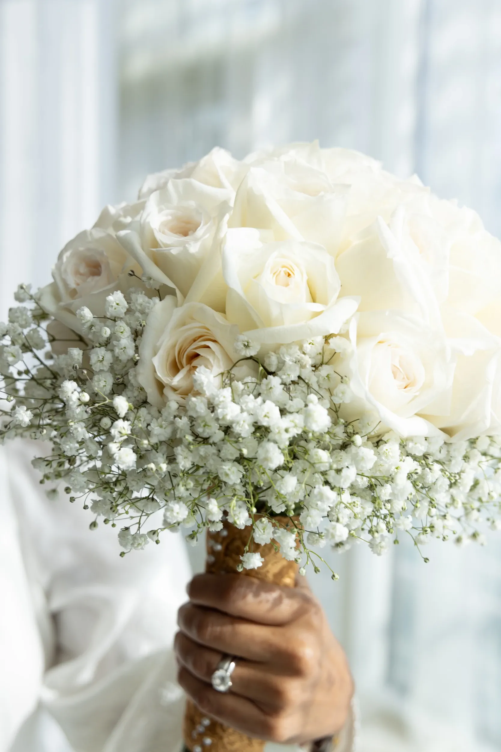 A white bridal bouquet held against softly lit curtains.