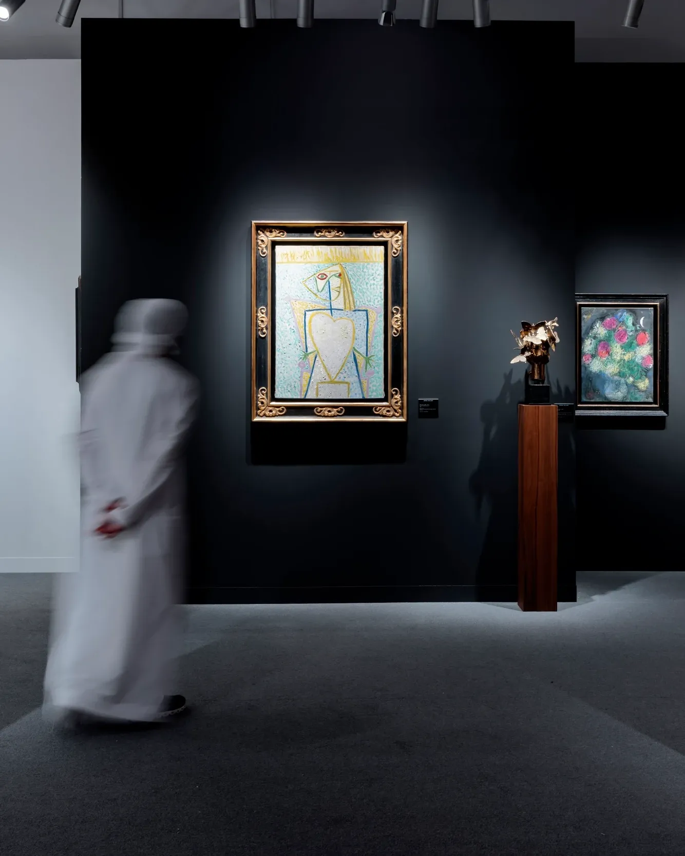 A blurred visitor in a kandura walking past a framed painting and a sculpture on a dark exhibition wall.